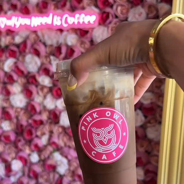 An iced coffee held up in front of the pink rose wall and the Owl You Need Is Coffee neon